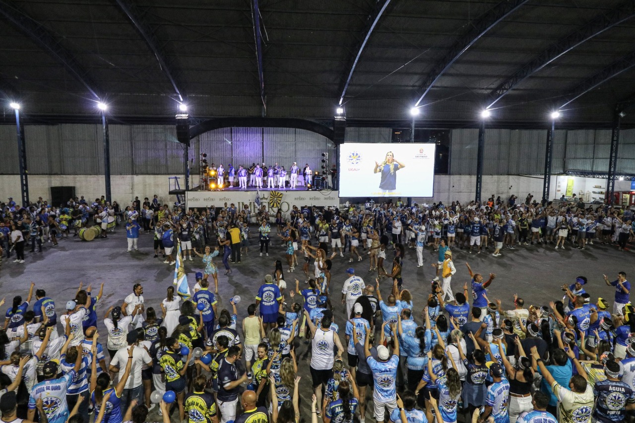 Na foto, na quadra da escola Águia de Ouro, é possível visualizar na primeira fileira as autoridades que participaram do lançamento do Samba com as Mãos, entre elas: o prefeito Ricardo Nunes e a secretária Silvia Grecco. Ao fundo, a quadra está lotada de integrantes da escola. Todos assistem a apresentação do Samba com as Mãos no telão.